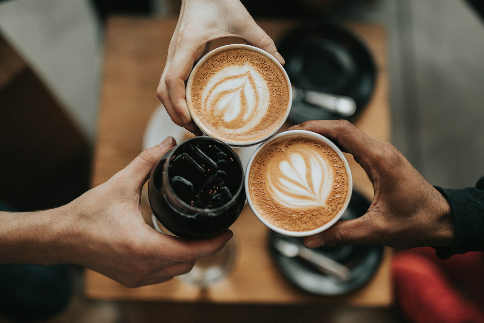 Three hands holding coffee cups together over a wooden table, two with latte art. It accompanies an article about Start AI automation with one boring workflow.