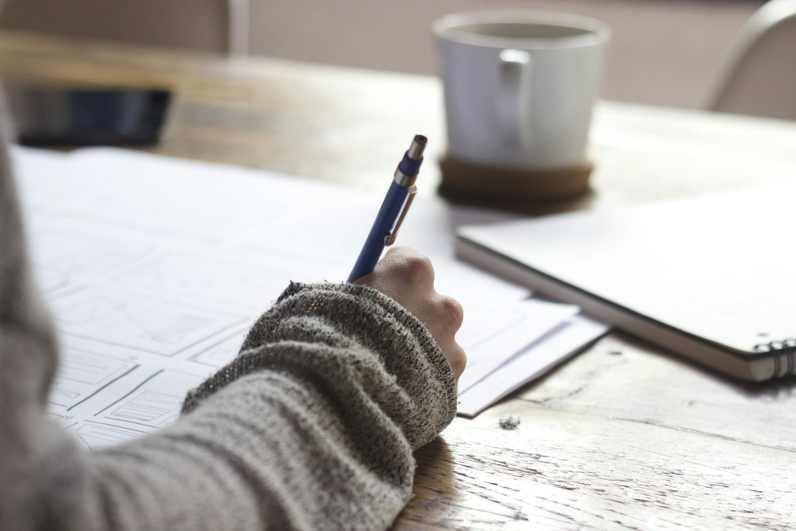 A hand holding a blue pen over wireframe sketches, with a mug and a closed notebook nearby. It accompanies an article about Scoping a project when the idea is still loose.