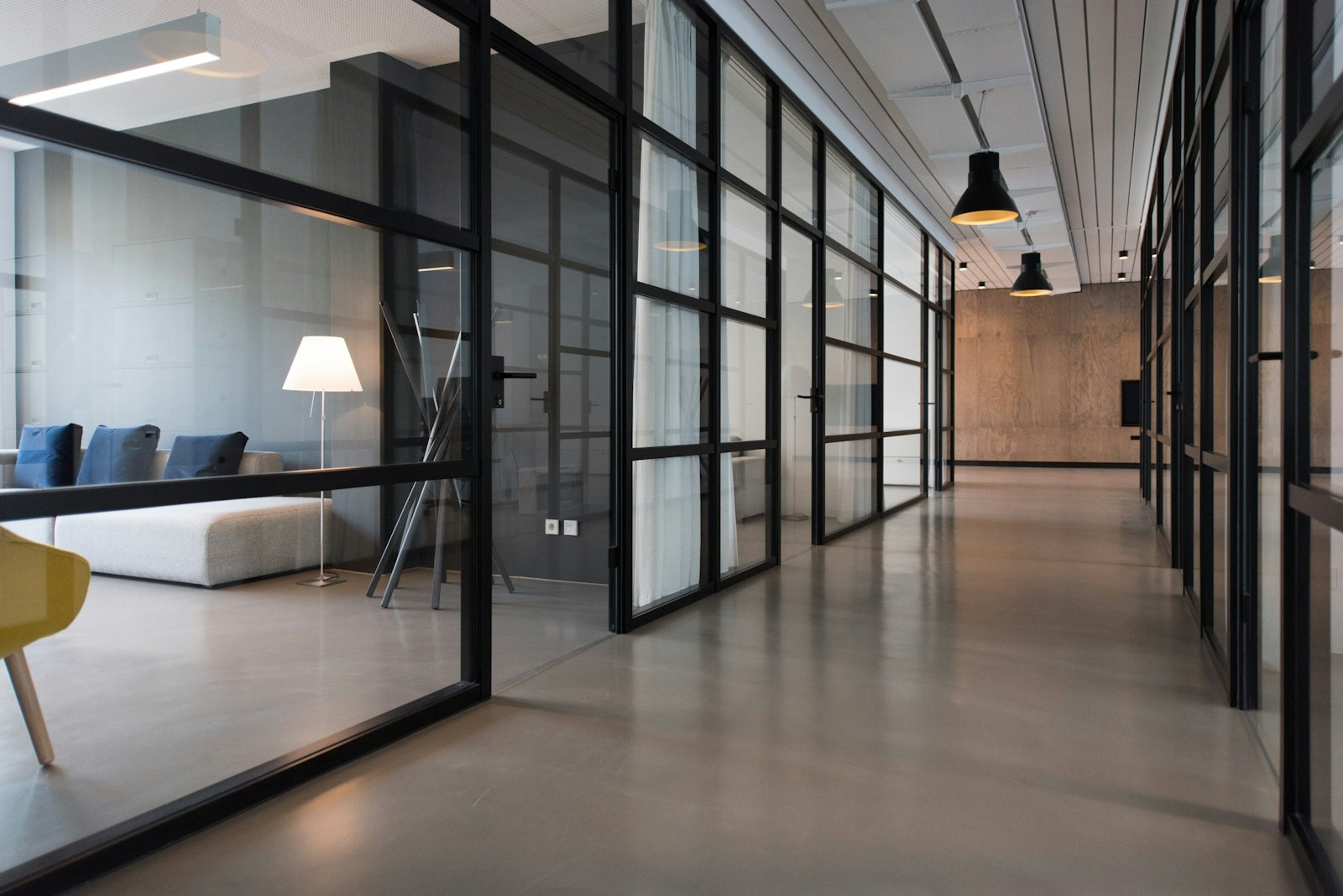A glass-walled office corridor with a sofa, a floor lamp, and black pendant lights. It accompanies an article about Choosing a CMS when the marketing team is small.